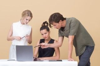 Man pointing to grey computer, with two women next to him.
